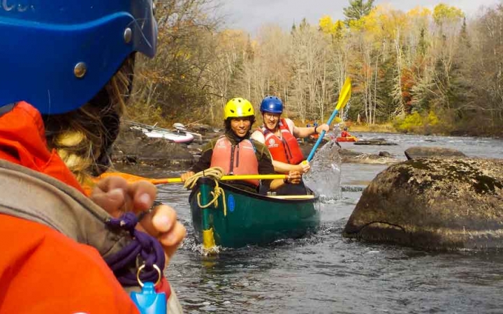 in the foreground, a person wearing whitewater gear looks on toward two people paddling a canoe on an outward bound expedition in maine